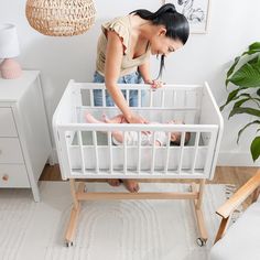 a woman standing over a baby in a crib next to a white dresser and potted plant