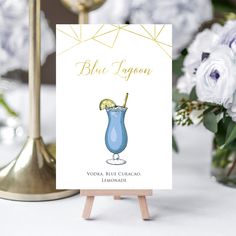 a card with a blue cocktail on it sitting next to a vase filled with flowers