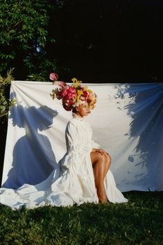 a woman sitting on top of a grass covered field next to a white sheet with flowers in her hair