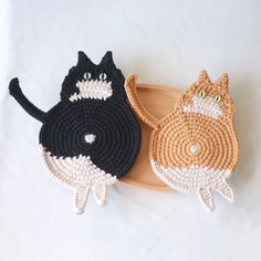 two crocheted cat coasters sitting on top of a wooden board with one black and the other orange