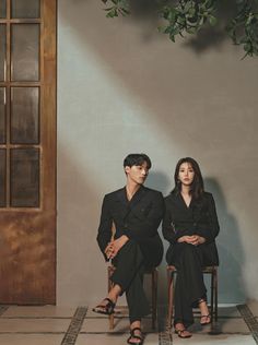 two people sitting on chairs in front of a wall with plants hanging from it's sides