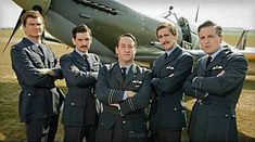 four men in uniforms standing next to an airplane with their arms crossed and looking at the camera