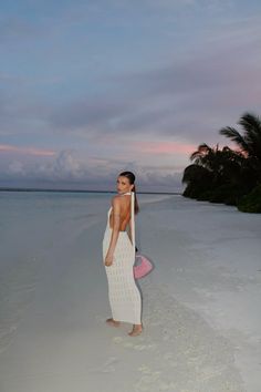 a woman standing on the beach at sunset with her back to the camera, wearing a white dress and carrying a pink purse