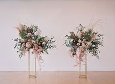 two tall vases with flowers and greenery in them on wooden floor next to wall