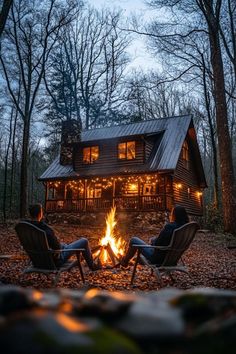 two people sitting in chairs around a campfire with the cabin lit up behind them