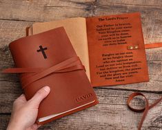 a hand holding a brown leather book with a cross on the front and an open bible behind it