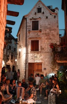 a group of people sitting around tables in front of an old building at night with lights on