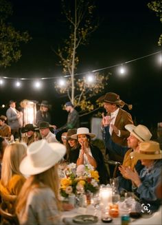 a group of people sitting around a table at a dinner party with lights strung overhead