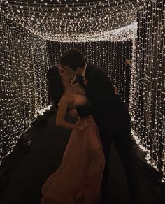 a man and woman are kissing in front of some string lights on the wall behind them