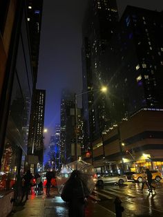 people with umbrellas are walking down the street in the rain at night, near tall buildings