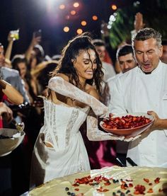 a woman in a white dress is holding a plate with strawberries on it while a man in a white suit and tie stands next to her