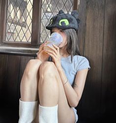 a woman sitting on the floor drinking from a bottle with a cat hat on her head