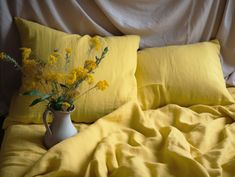 a white vase with yellow flowers sitting on a bed next to two pillows and a blanket