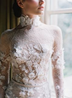 a woman wearing a white dress with flowers on it's neck and shoulders, standing in front of a window