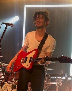 a young man holding a red guitar in front of a microphone and drum set on stage