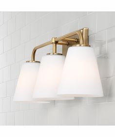 three light bathroom fixture with white glass shades on the bottom and gold finish, against a white tiled wall