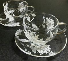 two clear cups and saucers with white flowers on them, sitting on a black surface