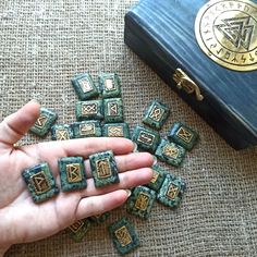 a hand is holding some small green and gold tiles in front of a black box