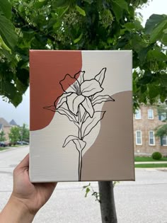 a hand holding up a piece of art that has flowers on it and is in front of a tree