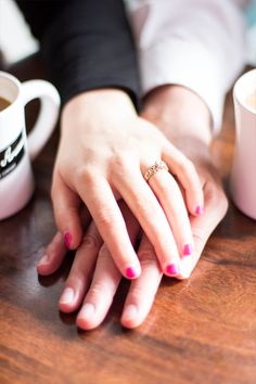 two people sitting at a table with their hands on each other's fingers, holding coffee mugs