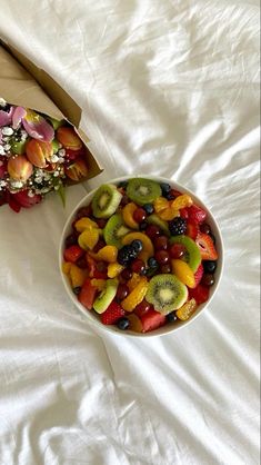 a bowl of fruit sits next to a box of flowers on a white bed sheet