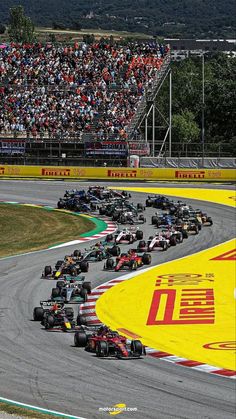 a group of cars driving down a race track with spectators in the stands behind them