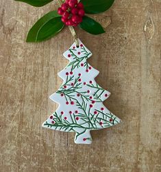 a ceramic christmas tree ornament hanging on a wooden table with holly and berries