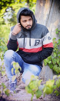 a man sitting on the ground next to a tree wearing a hoodie and jeans