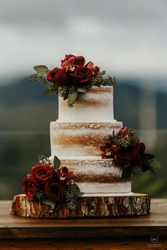 a three tiered wedding cake with red flowers on top is sitting on a wooden table