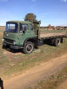 an old green truck parked on the side of a dirt road next to a field