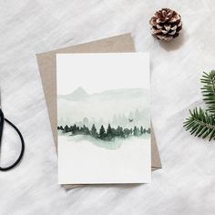 a card with a watercolor landscape on it next to scissors and pineconi