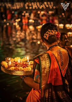 a woman holding a tray with candles on it in front of some water and lights