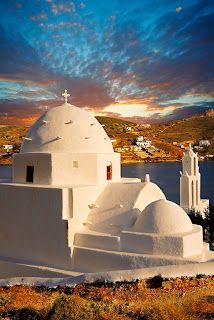 a white building with a cross on it's roof next to water and hills