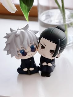 two anime figurines sitting next to each other on a table with flowers in the background