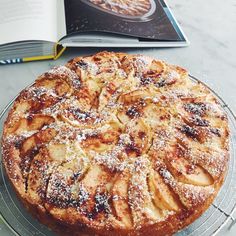 an apple pie with powdered sugar sits on a glass plate next to a cookbook