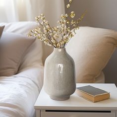 a white vase with some flowers in it on a table next to a book and pillows