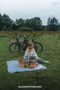 a woman sitting on a blanket in the grass next to two bikes and holding a picnic basket