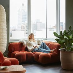 a woman sitting on top of a red couch in front of a window next to a potted plant