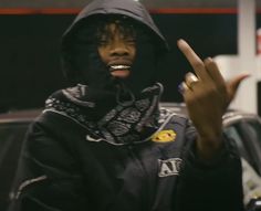 a man in a black hoodie making the peace sign with his hand while standing next to a car