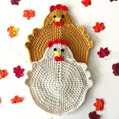 two crocheted roosters sitting next to each other on a white surface surrounded by fall leaves