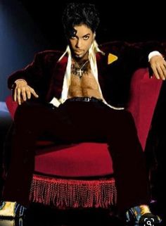 prince sitting in a red chair with his legs crossed and shirtless torso exposed, looking to the side