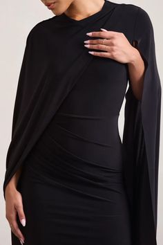 a woman wearing a black dress with an open cape over her shoulders and hands on her chest