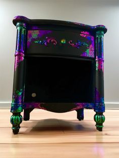 a black and purple nightstand with colorful paint on the top, sitting on a hard wood floor