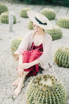 a woman sitting on the ground in front of cactus plants wearing a white hat and pink dress