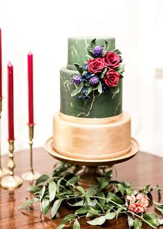 a three tiered cake with flowers and greenery sits on a table next to candles