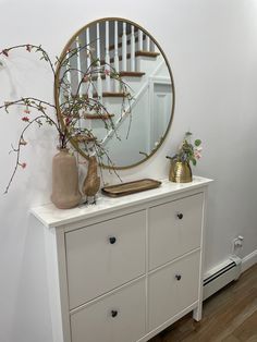 a white dresser topped with a round mirror and vase next to a wooden stair case