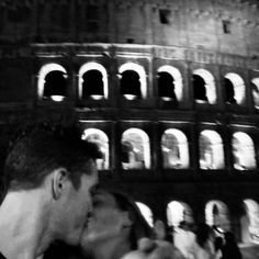 black and white photograph of two people kissing in front of the colossion at night