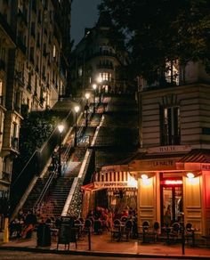 people are sitting at tables on the steps in front of buildings and stairs lit up by street lights