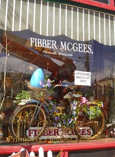 a store front window with flowers in baskets and an easter egg on the bike behind it