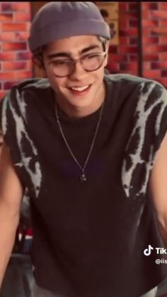 a young man wearing glasses and a black shirt smiles at the camera while sitting in front of a brick wall